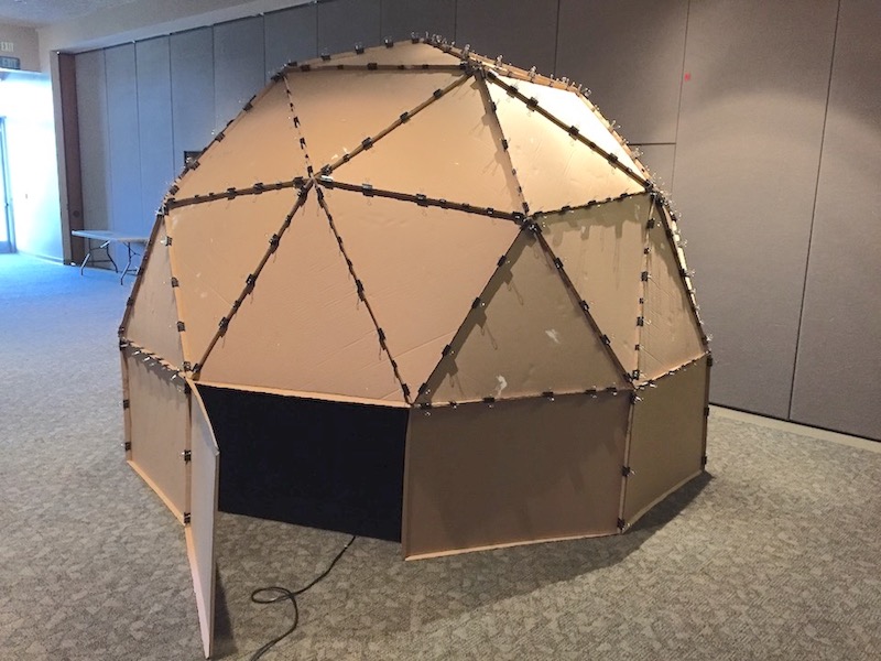 A 12-foot diameter cardboard geodesic dome, resting on a 2-foot cardboard base wall, with a door allowing people to enter